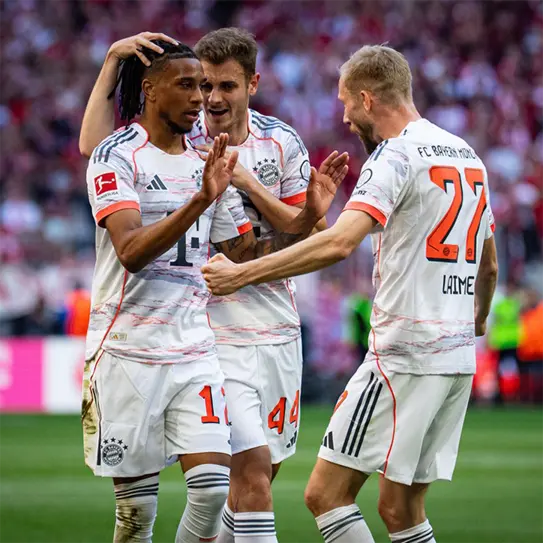 Three soccer players from Bayern Munich celebrate together after a successful play. One player, wearing jersey number 14, is at the center, smiling and gesturing. The other two players, in the background, show excitement and support. The atmosphere is energetic and celebratory.