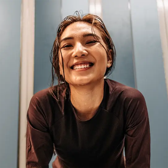 A smiling young woman with wet hair leans forward, exuding an energetic and cheerful vibe. She wears a dark athletic top and is set against a background of soft blue tones, suggesting a refreshing and active environment.