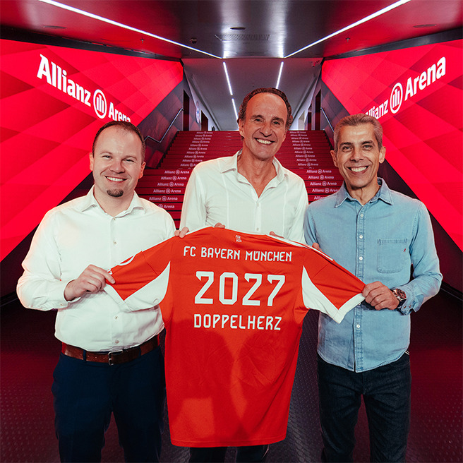 Three men stand together in a corridor at Allianz Arena, holding up a red FC Bayern München jersey. The jersey displays the number "2027" and the word "DOPPELHERTZ." The background features red lighting, enhancing the atmosphere of a sports event.