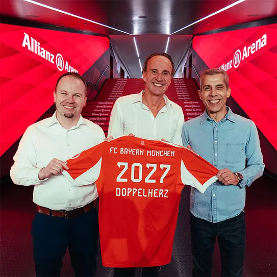 Three men stand together in a corridor at Allianz Arena, holding up a red FC Bayern München jersey. The jersey displays the number "2027" and the word "DOPPELHERTZ." The background features red lighting, enhancing the atmosphere of a sports event.