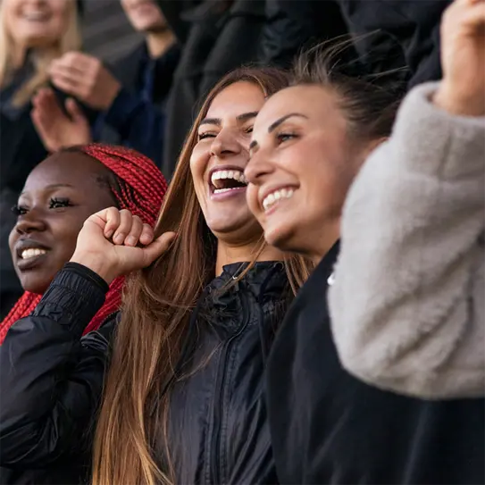Three women are smiling and laughing in a spirited group setting. They appear to be enjoying an event, with one woman waving her arm joyfully. The atmosphere is lively, conveying a sense of celebration and camaraderie.